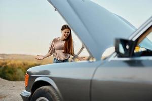 een verdrietig vrouw heeft geopend de kap van een gebroken naar beneden auto en is op zoek voor de oorzaak van de afbreken Aan een weg reis alleen met haar handen verspreiding deel en geschreeuw in woede foto