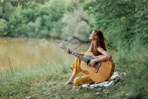 hippie vrouw spelen gitaar glimlachen en het zingen liedjes in natuur zittend Aan een plaid door de meer in de avond in de stralen van de instelling zon. een levensstijl in harmonie met de lichaam en natuur foto