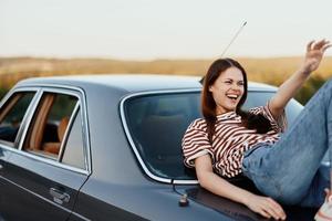 een jong vrouw zit Aan de romp van een auto, lacht en rust na een moeilijk weg en bewondert natuur met een mooi visie. stoppen is ook een deel van de reis foto