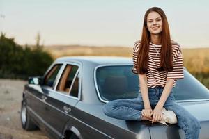 een jong vrouw zit Aan de romp van een auto en rust na een moeilijk weg en bewondert natuur met een mooi visie. stoppen is ook een deel van de reis foto