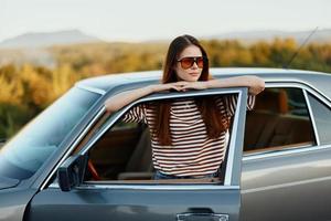 een jong vrouw bestuurder looks uit van de auto Bij de herfst landschap en glimlacht bevredigend foto