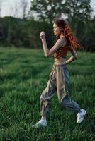 een vrouw met lang rood haar- werken uit en loopt Aan de groen gras in de park in joggingbroek en sportschoenen foto