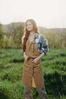 een boer vrouw in haar werk kleren, plaid overhemd en schort, staat in de veld- Aan de groen gras en glimlacht in de instelling zon na een moeilijk dag werk foto