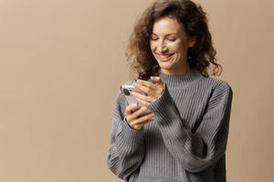 lief gelukkig gekruld mooi vrouw in grijs gewoontjes trui chatten met vriendje poseren geïsoleerd Aan over- beige pastel achtergrond. sociaal media, netwerk, afstand communicatie concept. kopiëren ruimte foto