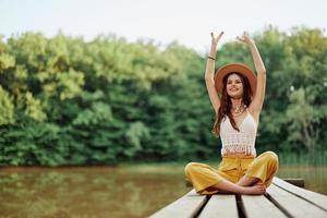 hippie eco-activist vrouw reiziger zit Aan een brug door een meer met haar armen uitgestrekt met een hoed en glimlachen vriendelijke groet foto
