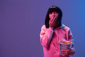 geschokt bang geweldig brunet vrouw in roze capuchon eten popcorn kijken terzijde poseren geïsoleerd in blauw paars kleur licht studio achtergrond. neon partij cyberpunk bioscoop concept. kopiëren ruimte foto