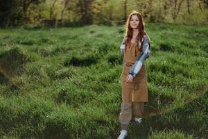 een vrouw tuinman in een schort staat in een veld- van groen gras buitenshuis, glimlachen Aan een zomer middag in een zonnig zonsondergang na een dag werk foto