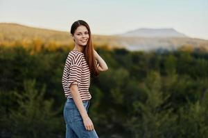 een jong vrouw lacht en looks Bij de camera in gemakkelijk kleren tegen de backdrop van een mooi landschap van bergen en bomen in herfst. levensstijl Aan de Actie foto