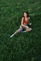 freelancer vrouw genieten van de buitenshuis zittend in de park Aan de groen gras in gewoontjes kleding met lang rood haar- lit door de helder zomer zon zonder muggen foto