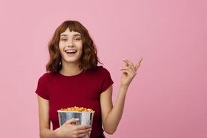 glimlachen vrolijk mooi schattig roodharige dame in rood overhemd met popcorn klaar naar kijk maar film poseren geïsoleerd Aan over- roze studio achtergrond. kopiëren ruimte spandoek. mode bioscoop concept. vermaak aanbod foto