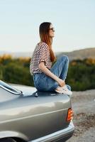 een mode vrouw in elegant bril, een gestreept t-shirt en jeans zit Aan de romp van een auto en looks Bij de mooi natuur van herfst foto