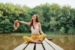 hippie eco-activist vrouw reiziger zit Aan een brug door een meer met haar armen uitgestrekt met een hoed en glimlachen vriendelijke groet foto