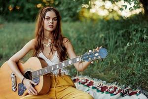 meisje hippie vrouw spelen gitaar in milieuvriendelijk kleren zittend Aan de grond buiten in natuur in de vallen aan het kijken de zonsondergang foto