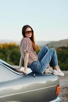 een mode vrouw in elegant bril, een gestreept t-shirt en jeans zit Aan de romp van een auto en looks Bij de mooi natuur van herfst foto