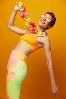 vrouw met bloemen hawaiiaans krans in de omgeving van haar nek heeft pret dansen en glimlachen in helder kleren Aan oranje achtergrond met zonnebril, levensstijl partij in hawaiiaans stijl foto