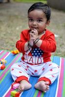 schattig weinig Indisch zuigeling zittend genieten van buitenshuis schieten Bij maatschappij park in Delhi, schattig baby jongen zittend Aan kleurrijk mat met gras in de omgeving van, baby jongen buitenshuis schieten foto