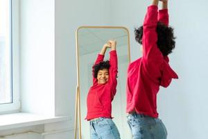 liefde jezelf. mooi jong glimlachen Afrikaanse Amerikaans vrouw dansen genieten van haar spiegel reflectie. zwart dame op zoek Bij spiegel op zoek zelfverzekerd en gelukkig. zelf liefde concept. foto