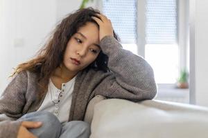 ongelukkig Aziatisch vrouw Aan sofa huilen. eenzaam verdrietig vrouw diep in gedachten zittend dagdromen of aan het wachten voor iemand in de leven kamer met een echt uitdrukking, zittend Aan bankstel foto