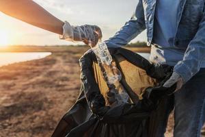 mensen vrijwilliger houden vuilnis plastic fles in zwart zak Bij park in de buurt rivier- in zonsondergang foto