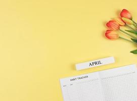 vlak lay-out van gewoonte tracker boek, houten kalender april, en tulpen Aan geel achtergrond met kopiëren ruimte. foto