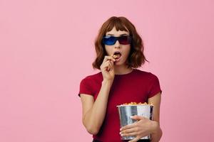 verward geconcentreerd schattig roodharige dame in rood t-shirt 3d bril eten popcorn kijk maar film poseren geïsoleerd Aan over- roze studio achtergrond. kopiëren ruimte spandoek. mode bioscoop concept. vermaak aanbod foto