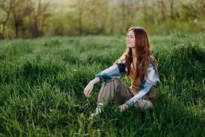 een jong mooi vrouw zit Aan de groen gras in de park en looks uit in de instelling zon foto