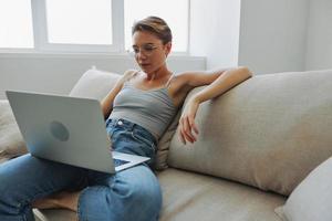 tiener- meisje freelancer met laptop zittend Aan bankstel Bij huis glimlachen in huis kleren en bril met kort kapsel, levensstijl met Nee filters, vrij kopiëren ruimte foto