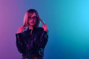 flirten sexy aanbiddelijk mooi blond vrouw in leer jasje sparkly jurk modieus zonnebril poseren geïsoleerd in blauw roze kleur licht studio achtergrond. neon partij cyberpunk concept. kopiëren ruimte foto