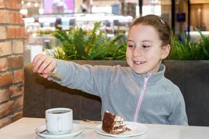 glimlachen weinig meisje in een cafe Bij de tafel giet suiker in een kop De volgende naar een bord met taart foto