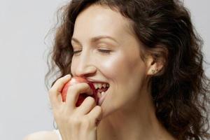 detailopname portret van gelukkig gekruld mooi vrouw in eenvoudig wit t-shirt genieten vers appel fruit poseren geïsoleerd Aan over- wit achtergrond. natuurlijk milieuvriendelijk producten concept. kopiëren ruimte foto