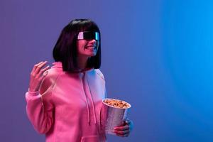 lachend geweldig brunet vrouw in roze capuchon modieus spiegelend zonnebril met popcorn verspreiding hand- kijken terzijde poseren geïsoleerd in blauw paars kleur licht achtergrond. neon partij bioscoop concept. kopiëren ruimte foto