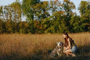 een vrouw Toneelstukken en dansen met een schor ras hond in natuur in herfst Aan een veld- van gras en glimlacht Bij een mooi zo avond in de instelling zon foto