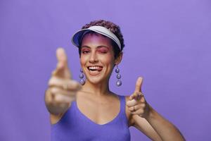 portret van een sportief mode vrouw poseren glimlachen met tanden en richten een vinger Bij de camera in een Purper yoga trainingspak en een transparant pet Aan een Purper monochroom achtergrond foto