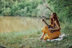 een hippie vrouw spelen haar gitaar glimlacht en zingt liedjes in natuur zittend Aan een plaid in de avond in de zonsondergang zonlicht. een levensstijl in harmonie met de lichaam en natuur foto