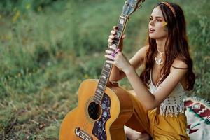 gelukkig hippie vrouw met een gitaar ontspannende in natuur zittend Aan een plaid glimlachen en genieten van de visie. levensstijl in harmonie met natuur en zelf foto