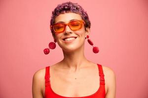 mode portret van een vrouw met een kort kapsel in gekleurde zonnebril met ongebruikelijk accessoires met oorbellen glimlachen Aan een roze helder achtergrond foto