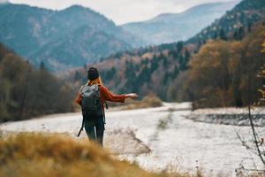 vrouw reiziger met een rugzak en in een hoed in de buurt de rivier- in de bergen in herfst foto