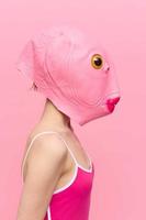 een vrouw in een roze vis hoofd masker staat in profiel tegen een roze achtergrond en looks in de camera met een geel oog, een gek conceptuele halloween kostuum foto
