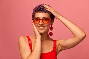 mode portret van een vrouw met een kort kapsel in gekleurde zonnebril met ongebruikelijk accessoires met oorbellen glimlachen Aan een roze helder achtergrond foto