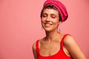 jong atletisch vrouw met een kort kapsel en Purper haar- in een rood top en een roze hoed met een atletisch figuur glimlacht en grimassen op zoek Bij de camera Aan een roze achtergrond foto