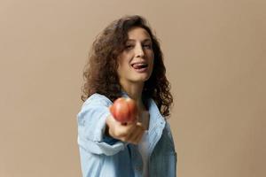 schattig vrolijk gelukkig gekruld mooi vrouw in jeans gewoontjes overhemd trekt appel Bij camera poseren geïsoleerd Aan over- beige pastel achtergrond. gezond voedsel. natuurlijk milieuvriendelijk producten concept. kopiëren ruimte foto