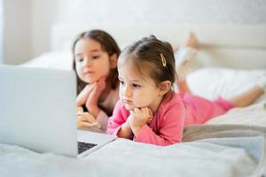 twee schattig weinig zussen slijtage pyjama op zoek tekenfilm door laptop in ochtend- Bij huis. foto