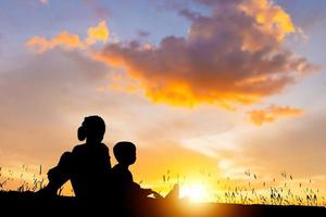 silhouet van jongen en meisje met gitaar hebben pret buitenshuis, portret van aanbiddelijk broer en zus spelen buitenshuis. Aziatisch kinderen het zingen liedjes met zonsondergang achtergrond foto