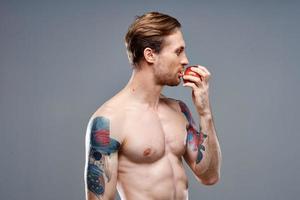 sexy atleet met gemotiveerd omhoog spieren eet een appel Aan een grijs achtergrond en een tatoeëren Aan zijn arm foto