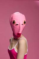 een vrouw vervelend een roze vis hoofd masker Aan halloween staat en looks Bij de camera tegen een roze achtergrond. conceptuele kunst foto in een gek manier