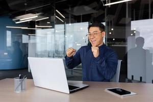 geslaagd zakenman vieren zege, Mens dansen zittend Bij tafel, Aziatisch Mens in overhemd en bril Bij werk binnen kantoor met laptop, tevreden met prestatie investeerder foto