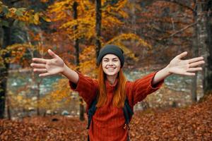 gelukkig vrouw met een rugzak reist in de park in herfst en gebaren met de handen van de rivier- in de achtergrond foto