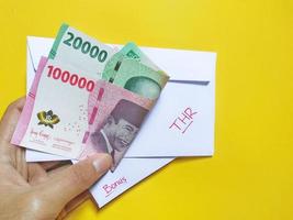 Mens Holding een wit envelop geschreven van door, bonus en nieuw Indonesisch bankbiljetten, meestal tunjangan hari raya of gebeld thr zijn gegeven naar medewerkers verder van eid. geïsoleerd Aan geel achtergrond foto