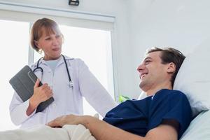 de dokter is onderzoeken de geduldig in de ziekenhuis. Kaukasisch vrouw dokter pratend naar mannetje geduldig aan het liegen in ziekenhuis bed. foto