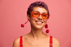 mode portret van een vrouw met een kort kapsel in gekleurde zonnebril met ongebruikelijk accessoires met oorbellen glimlachen Aan een roze helder achtergrond foto